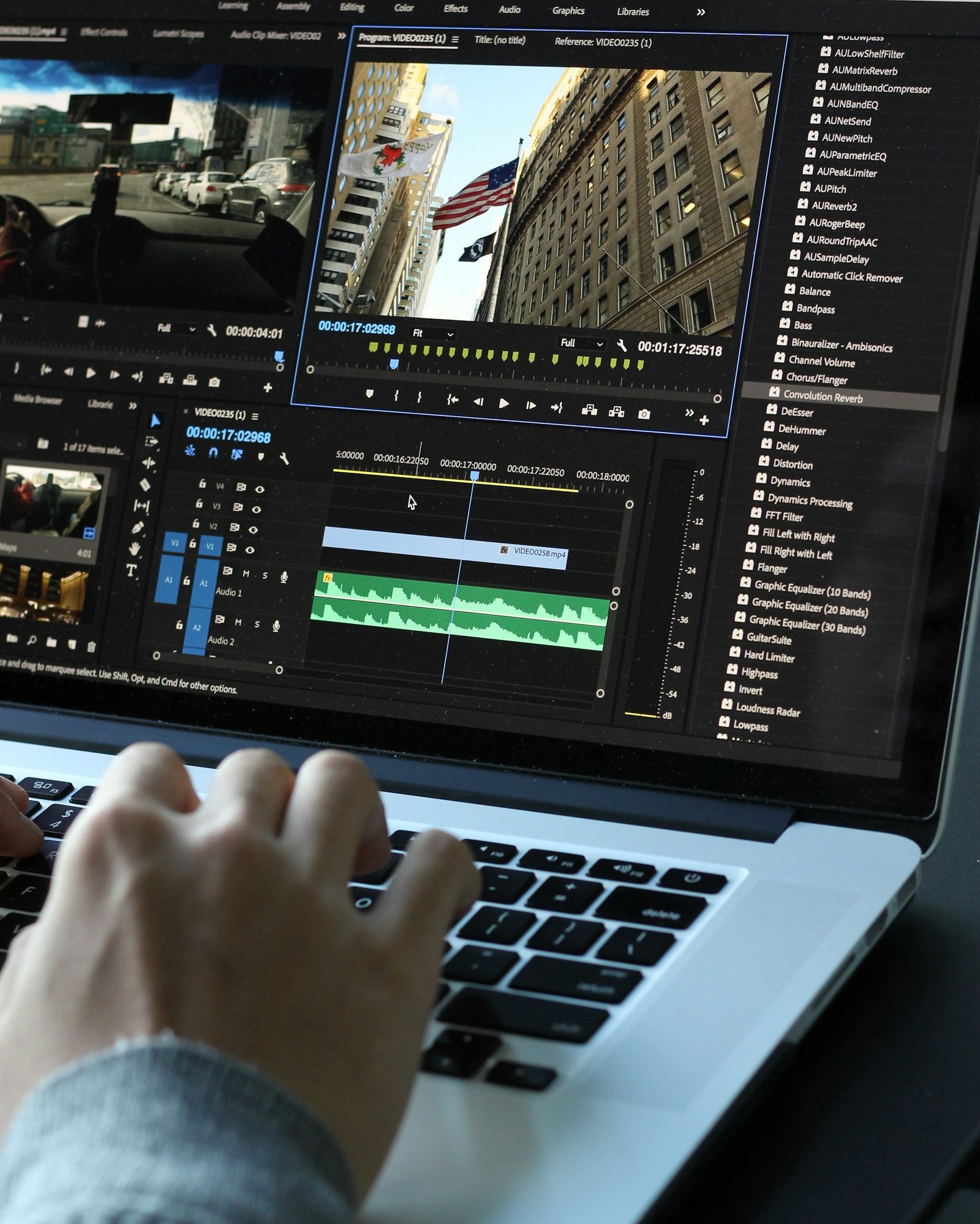 Hands typing on a laptop running Premiere Pro, with a timeline and audio waveforms on screen